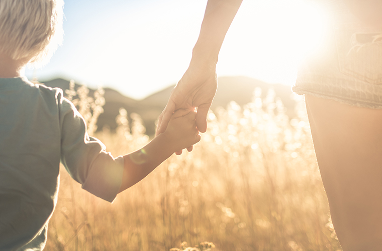 Mother little child holding hands walking in a grass field at sunset