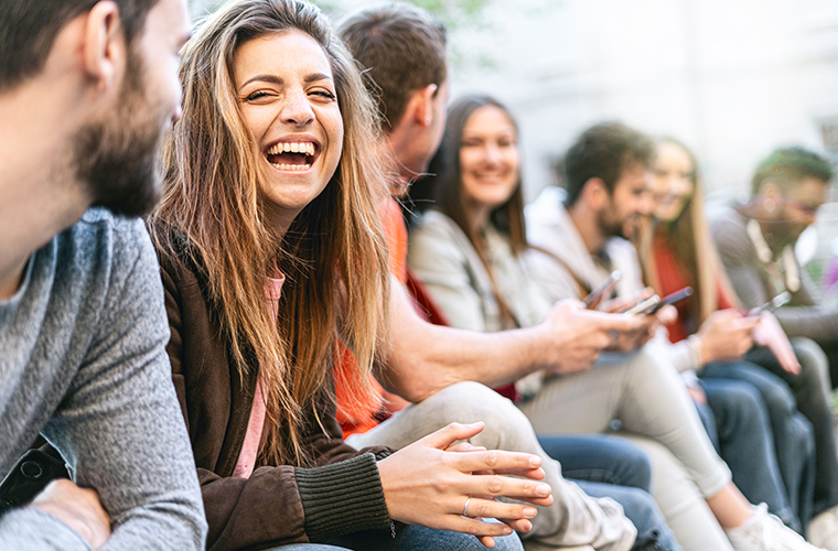 Group of trendy young people chatting together sitting on a bench outdoors. Students having fun together. Focus on a blonde girl smiling
